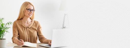 A woman sits at a light, airy workspace, focused on her laptop while taking notes in a notebook. A potted plant adds a touch of greenery, enhancing the cozy atmosphere, copy space photo