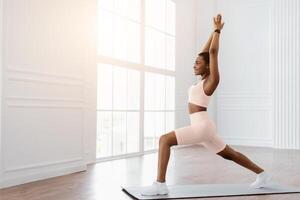 A millennial woman performs a lunging exercise in a sunny home environment. She focuses on maintaining her form while promoting her fitness routine and healthy lifestyle, copy space photo