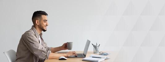 People And Technology. Profile portrait of young smiling Arab man using pc sitting at desk, typing on keyboard. Cheerful guy browsing internet, surfing web, holding cup of hot coffee, free copy space photo