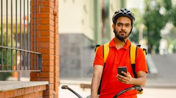 Home delivery service. Friendly courier in protective helmet, with yellow backpack, sits on bicycle and holds smartphone in hand on street, copy space photo