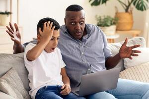 A man and a boy sit together on a couch, both displaying shock and excitement while looking at a laptop. Their expressions reflect a surprising and engaging experience taking place. photo