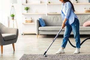 A young woman enjoys her time cleaning the carpet in her modern living room. She uses a vacuum cleaner while wearing casual clothes, with sunlight streaming through the window, copy space photo