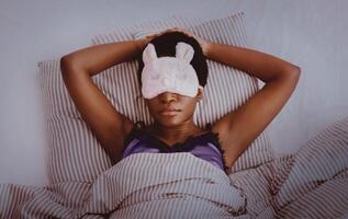 Cozy bed at night, sweet dream, rest at home and healthy lifestyle. Millennial african american female with funny mask, sleeping on bed in interior of bedroom, dark, top view, panorama, empty space photo