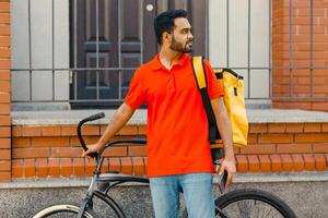 Work in city. Serious delivery man with beard with big yellow backpack and bicycle, holding smartphone and looking for address of client, outdoors photo