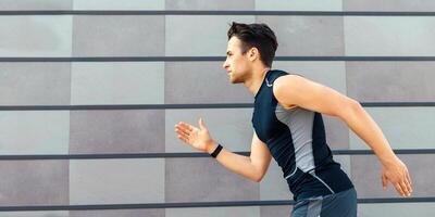 Jogging in morning. Handsome guy in sportswear and with fitness tracker, runs near gray wall background, free space photo