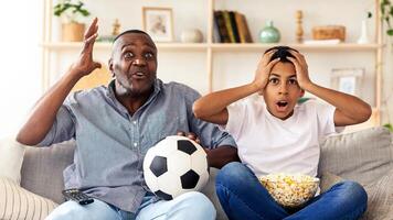 A man and a boy sit on a couch, transformed by the thrill of a soccer game. The boy looks shocked, while the man expresses enthusiasm, holding a soccer ball beside him. photo