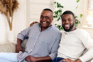 A father and son share a joyful moment together in their cozy living room, smiling warmly as they relax on the sofa surrounded by plants and comfortable decor, creating a heartwarming atmosphere. photo
