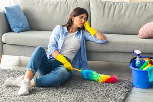 A young woman in casual attire sits on the floor of her living room, wearing yellow gloves. She looks bored while dusting the carpet with a colorful duster. Cleaning supplies are nearby. photo