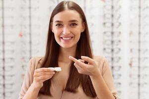A smiling woman holds a contact lens case and applies solution with care. The setting is bright and the background features various eyeglasses. photo