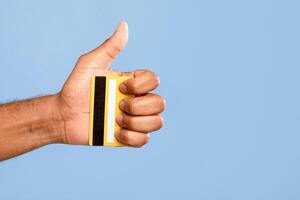 An African American man smiles and shows a thumbs up gesture while holding a yellow credit card in his hand. The background is a solid light blue, suggesting a positive and friendly vibe. photo