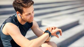 Modern jogging gadgets. Tired athlete with wireless headphones looks at fitness tracker and sits on steps, close up, free space, sun flare photo