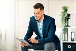 A young Middle Eastern man in a sleek suit interacts with a tablet while seated in a contemporary office. He appears focused and engaged in his work during daytime hours. photo