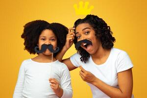 Funny african mother and daughter fooling together, playing with party accesories, having fun with paper moustaches on sticks over pink background photo