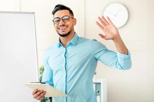 A young Arab man stands in a modern office environment. He is smiling and waving while holding a clipboard, ready to present ideas to colleagues. photo