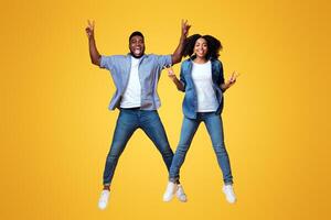 Two young adults, a man and a woman, are jumping in the air, smiling broadly. They are dressed casually in denim and look lively and carefree against a vibrant yellow backdrop. photo