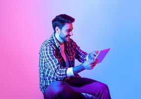 A man with a friendly smile interacts with a tablet while sitting comfortably. The backdrop features a blend of pink and blue hues, creating a lively atmosphere. photo