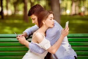 Gadget addicted couple using mobile phones while hugging on bench at park photo