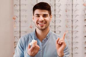 A young man wearing a blue shirt stands in an eyeglass store. He smiles and holds a contact lens case in his left hand. His right hand points up as if he is explaining something. photo