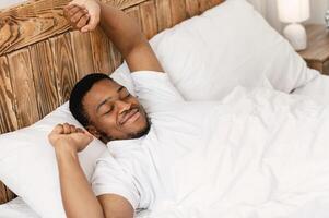 Good Morning. Rested Black Man Stretching Hands Waking Up Lying In Comfortable Bed At Home, Smiling With Eyes Closed. Wellslept Man Awakening After Good Sleep And Rest. High-Angle View photo