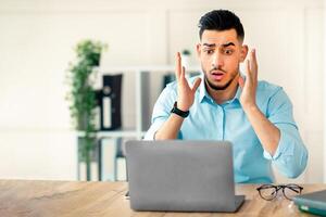 A young Arab man sits at a wooden desk in a modern office, visibly surprised by something on his laptop screen. He shows a mix of concern and disbelief, dressed in a casual button-up shirt. photo
