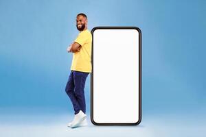 A man with a joyful expression stands confidently next to a large, blank smartphone. He wears a bright yellow shirt and casual pants, showcasing a modern lifestyle in a vibrant studio setting. photo