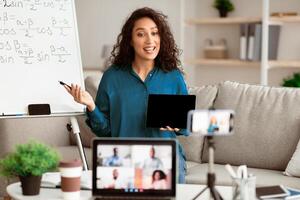 A young Middle Eastern woman engages students during an online tutoring session. She holds a tablet while demonstrating lessons on a board, creating an interactive learning experience. photo