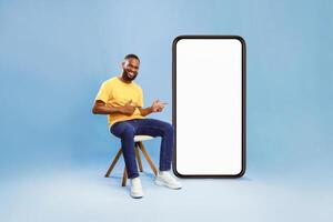 A millennial African American man smiles while seated on a wooden chair beside an oversized smartphone. The bright background highlights his relaxed lifestyle and the device's features. photo