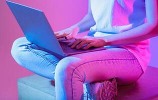 Woman sits on a block, typing on a laptop. The ambient lighting casts vibrant colors, creating a modern workspace vibe. Focus is on the hands and the laptop screen, croped photo
