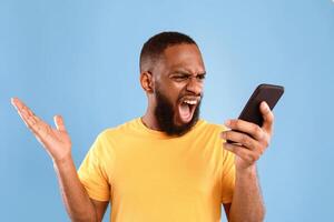A young African American man in a yellow shirt expresses frustration while looking at his smartphone against a blue background. photo
