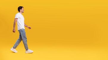 Full length body size profile side view of happy excited bearded guy in casual white t-shirt and jeans walking looking aside at free empty space isolated over orange color background, panoramic banner photo