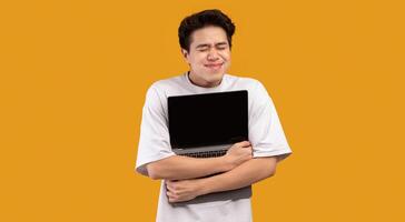 Technology Lover Concept. Happy excited asian man hugging laptop with black blank empty screen, holding it tight near chest. Geek posing isolated over orange studio background wall, copy space photo