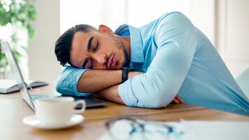A young Arab man is resting with his head on his arms at a desk in a modern office. The atmosphere indicates a long working day, with a laptop and paperwork nearby. photo