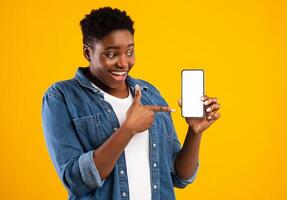 Cheerful Black Lady Showing Smartphone Blank Screen Recommending Application Posing Standing Over Yellow Studio Background. Mobile Application Ad. Check This App Concept. Mockup photo