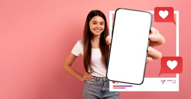A woman stands against a pink background, smiling while holding up a smartphone. The device displays a blank screen, ideal for showcasing social media applications and engagement. photo