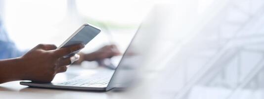Unrecognizable black woman using smartphone and working on laptop in white office, checking new mobile app, messaging on cellphone, browsing online banking, cropped image with selective focus photo