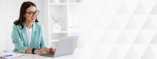 Cheerful millennial caucasian lady in suit and glasses typing on laptop at workplace in office interior. Business with gadget, communication with client remotely, manager work photo