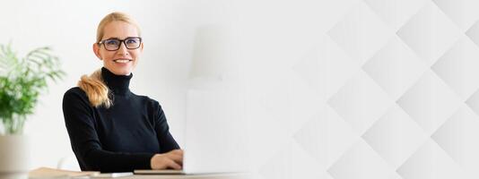 A woman with glasses smiles while sitting at her desk, engaging with a laptop. The modern office features greenery and a neutral color palette, creating a bright and inviting atmosphere, copy space photo
