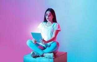 Asian woman sits cross-legged on a cube in a creative space, expressing surprise while using a tablet. The vibrant blue and pink lighting adds a dynamic atmosphere to the moment. photo