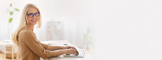 A woman with long hair and glasses sits at a desk, using a laptop. The workspace features natural light and indoor plants, creating a productive and inviting atmosphere for remote work, copy space photo