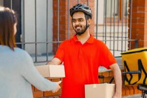 Fast eco service and delivery of parcels to home. Indian smiling millennial man in orange t-shirt and safety helmet gives to lady client a box with parcel, next to bicycle with yellow thermo bag photo
