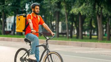 Modern work and delivery on eco transport. Young friendly indian man in uniform, protective gear with backpack riding on bicycle on sidewalk down the street, side view, panorama, empty space, outdoor photo