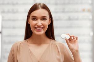 A young woman smiles while holding a contact lens case in an eyewear store. The store has various eyeglasses displayed in the background, indicating a vibrant shopping atmosphere. photo