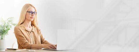 A focused woman with blonde hair types on her laptop in a well lit office space. She wears glasses and is surrounded by greenery, creating a calm and productive atmosphere, copy space photo
