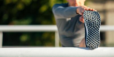 Flexibility and exercise. Muscular guy in sportswear puts his foot on railing and does stretching or exercise at park, close up, free space photo