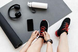 Preparing for morning workout at home or gym. Lady athlete ties shoelaces on sneakers, sits on floor with mat, bottle of water, headphones and smartphone with blank screen, cropped, studio shot photo