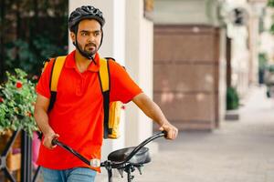 Business, work and delivery. Guy with beard in protective helmet with yellow backpack and bicycle walks down the street to client, close up, panorama, free space photo