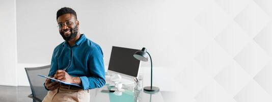 In a contemporary office space, a young African American businessman sits confidently with a clipboard. He smiles while showcasing a professional and focused approach to his work, copy space photo