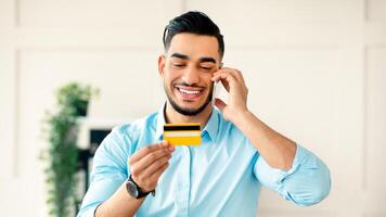 A young Arab entrepreneur is smiling while holding a credit card in one hand and talking on the phone. He appears engaged and focused in a bright office environment during work hours. photo