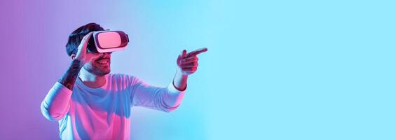 An enthusiastic man wears virtual reality goggles while pointing excitedly in a colorful setting. His expression shows immersion in a digital world, highlighting excitement and curiosity, copy space photo