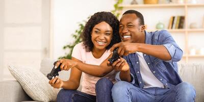 Weekend at home. Emotional african american man and woman with joysticks sitting on couch in living room, playing games and laughing, having fun together at home during lockdown, copy space photo